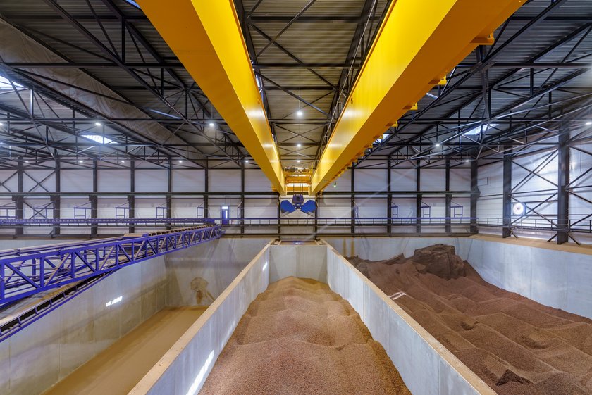 [Translate to Deutsch:] A warehouse from the inside. Three large compartments with cocoa can be seen. They are surrounded by blue and yellow conveyor belts and metal struts. An open blue shovel can be seen above the middle compartment.