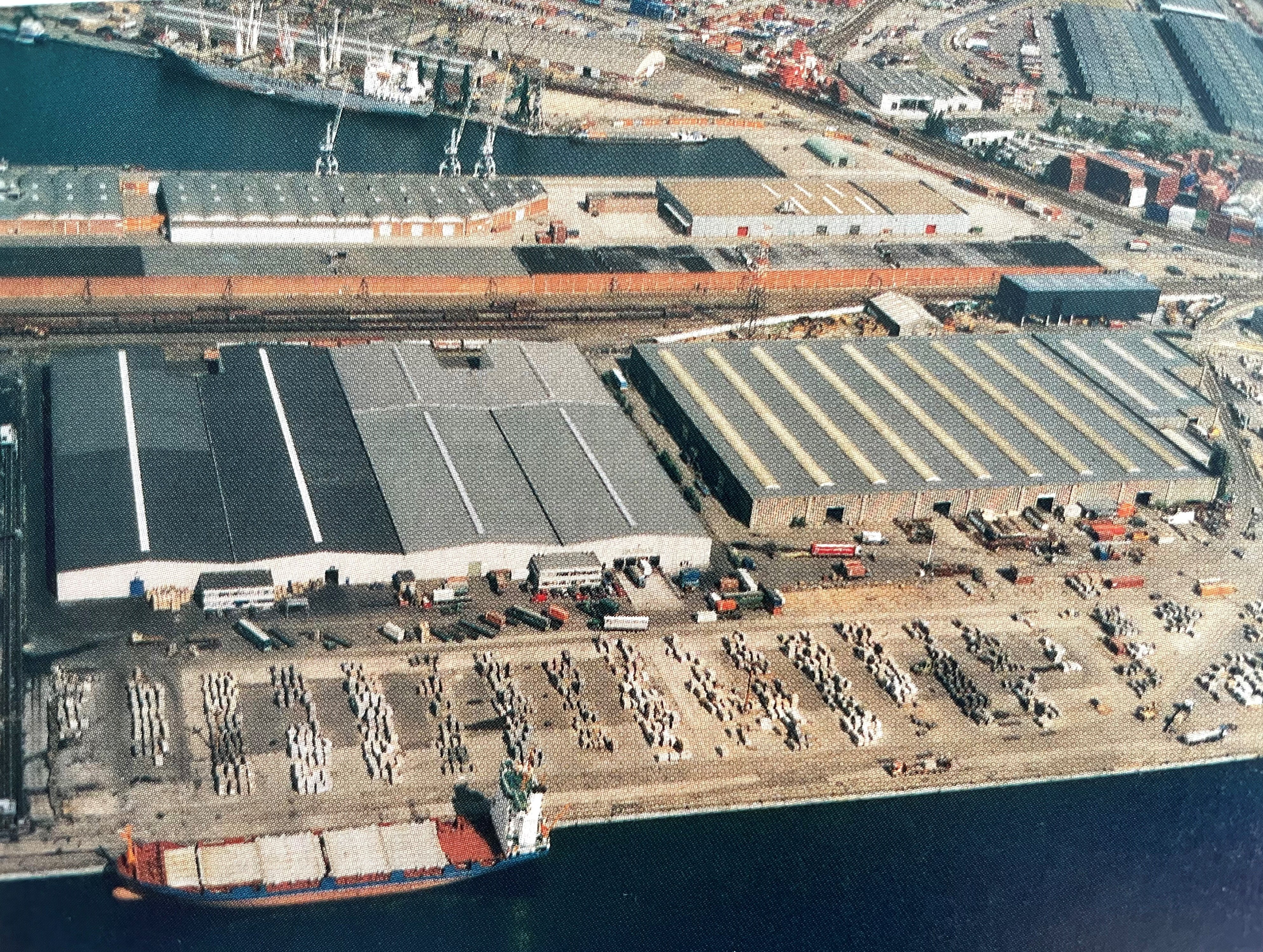 The photo is taken from above. You can see some water and a ship with containers in the front. Behind there are cars and trucks. In the center are warehouses. The back contains of buildings, possibly offices, followed by more water.