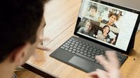 An opened notebook on a table. A video conference is held, four people are on the screen. One person is seen from behind, looking at the notebook.