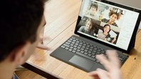 Ein geöffnetes Notebook steht auf einem Tisch. Eine Videokonferenz findet statt, vier Personen sind auf dem Bildschirm zu sehen. Eine Person ist von hinten zu sehen und schaut auf das Notebook.