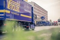 Close up of a blue truck with the yellow Vollers logo on it, in front of office buildings
