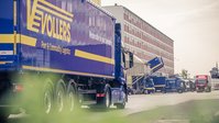 Close up of a blue truck with the yellow Vollers logo on it, in front of office buildings