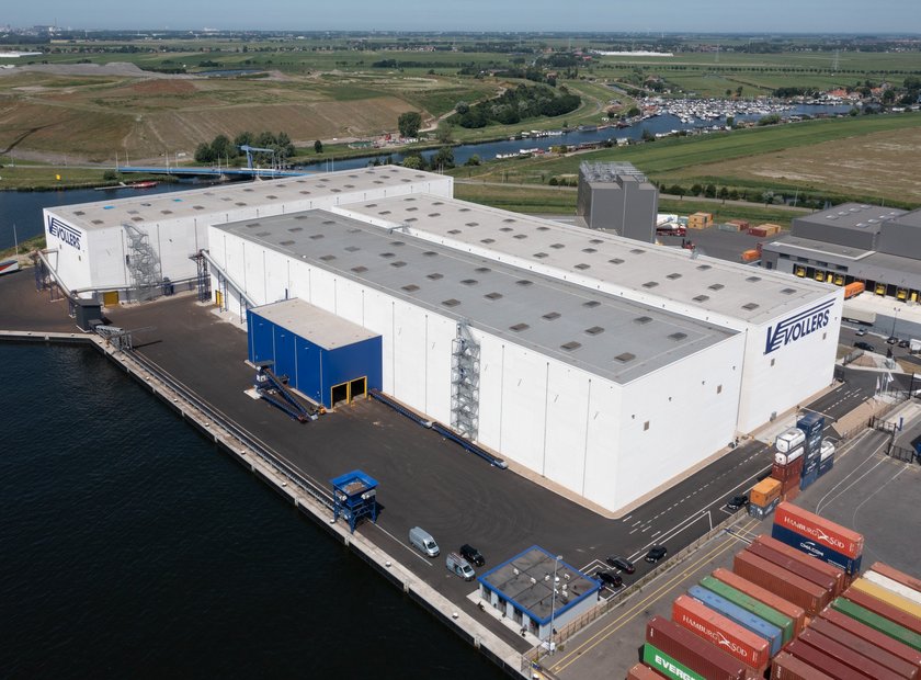 [Translate to Deutsch:] A photo taken from above. Centered are three white warehouses with a blue logo on the walls. It reads "Vollers". Left to the warehouses and behind it, is water. To the right, containers in all colours can be seen.