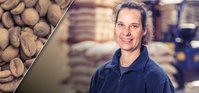 Woman with brown hair smiling at the camera in a warehouse with coffee bags and a forklift in the background.