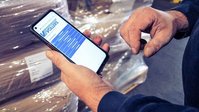 Person scannt einen Barcode auf gestapelter Fracht mit einem Smartphone in einer Lagerhalle.