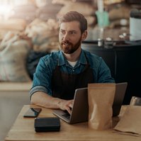 Ein Mann sitzt an einem Tisch mit einem Laptop, umgeben von Kaffeeproben in einer Lagerumgebung.