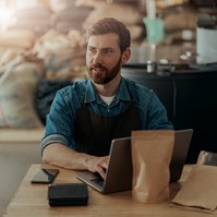 Een man zit aan een tafel met een laptop, omringd door koffiemonsters in een magazijnomgeving.