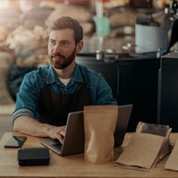 A man sitting at a table with a laptop, surrounded by coffee sample bags in a warehouse setting.