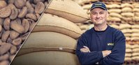 Man wearing a dark blue cap and sweater leaning against coffee bags in a warehouse, smiling at the camera.