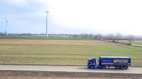Vollers-vrachtwagen rijdt over een weg in een landelijk landschap met windturbines op de achtergrond.