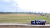 Vollers-Lkw fährt auf einer Straße durch eine ländliche Landschaft mit Windkraftanlagen im Hintergrund.