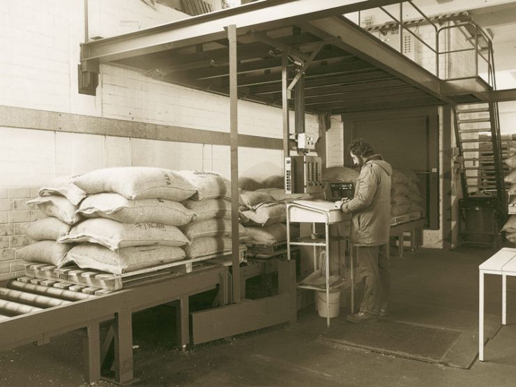 Historic sepia photo of workers handling coffee bags on a conveyor system in a warehouse.