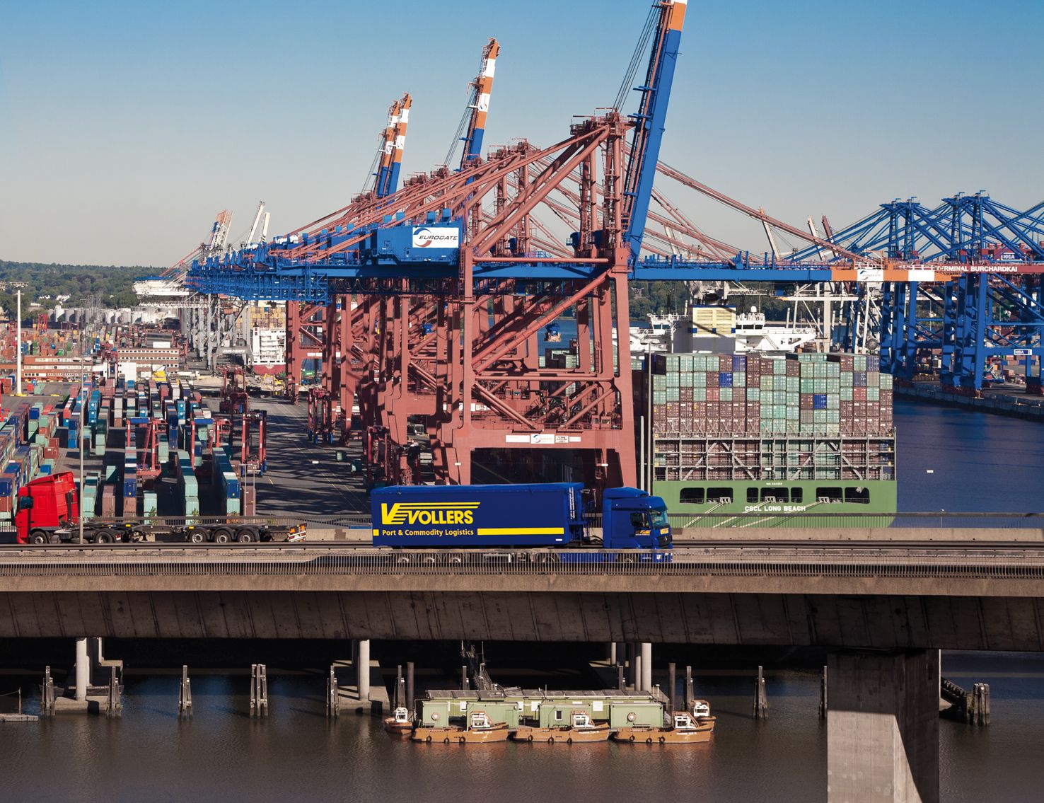 The truck port of Hamburg. A blue Vollers truck can be seen at the center of the photo. Behind it there are lots of colourful containers. Water can be seen in front and to the right of the truck.